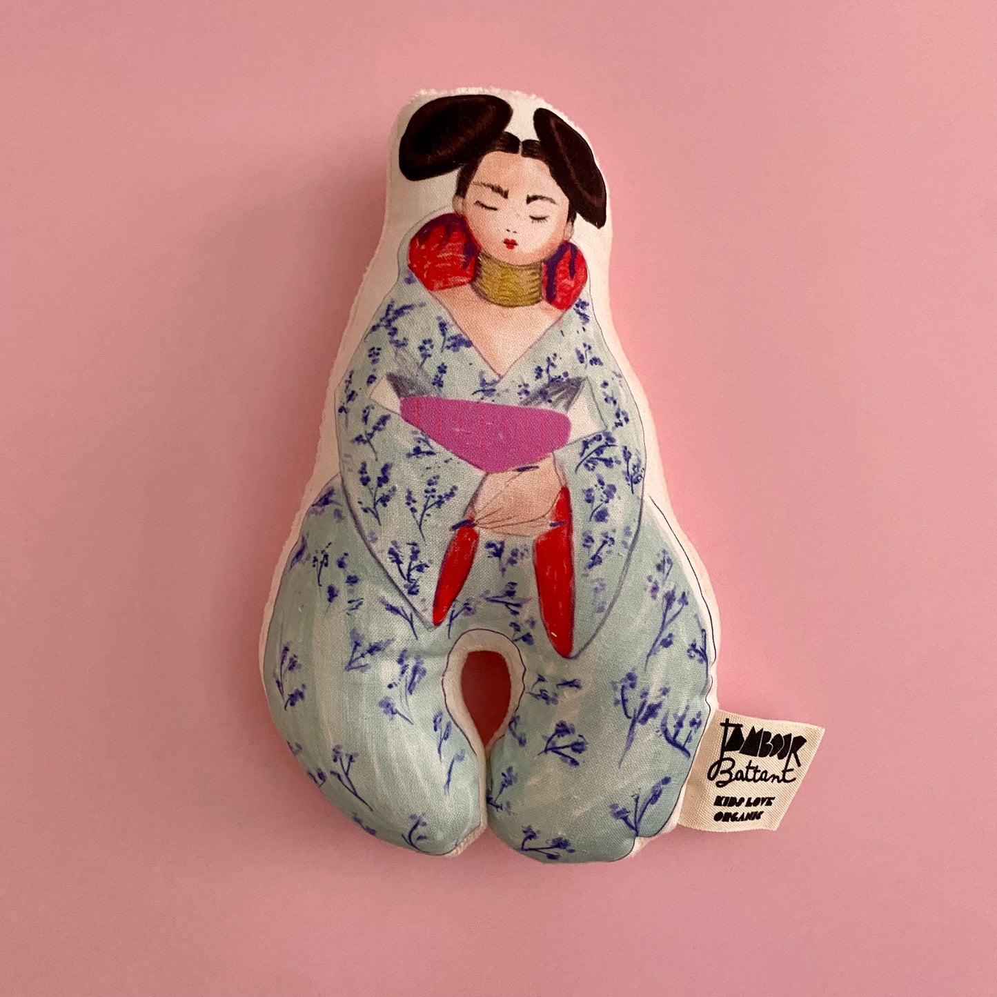 poupée hochet en coton bio faisant penser à une geisha, hommage à la chanteuse Björk. Poupée dessinée à l'aquarelle . La poupée est posée sur un fond rose uni.